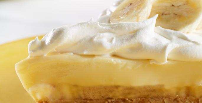 Fotografia de uma fatia de torta de mousse de banana, merengue e rodelas de banana por cima sobre um prato amarelo.