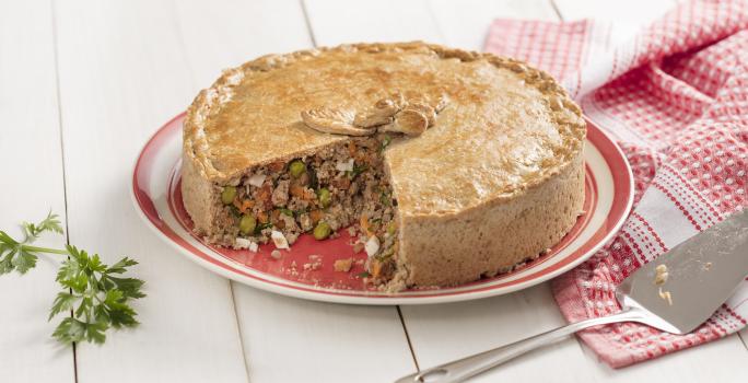 Fotografia em tons de branco, dourado e vermelho, com torta de carne assada e fatiada sobre prato vermelho e branco, ao lado guardanapo em branco e vermelho e uma espátula, ramo de salsa ao lado esquerdo, tudo sobre bancada de madeira na cor branca.