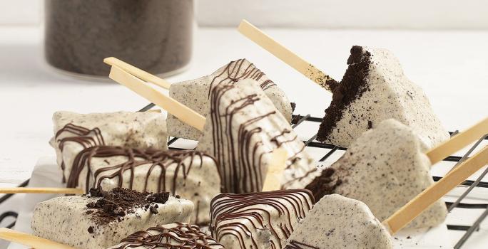 Fotografia em tons de branco em uma bancada de cor branca. No centro, estão os Cakes Pop Cookies’n Cream polvilhados com chocolate. Ao fundo, um recipiente contendo Topping de Negresco.