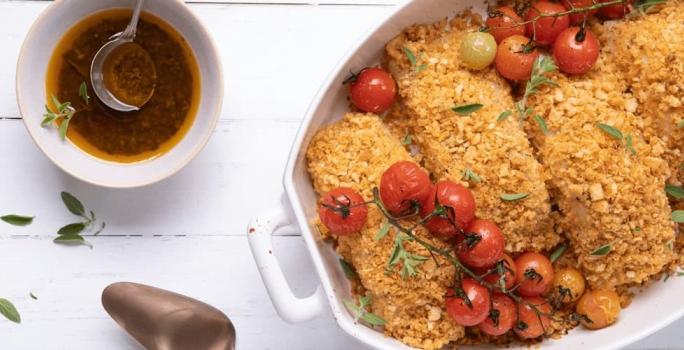 Fotografia em tons de amarelo em uma bancada de madeira branca com um refratário oval branco com as postas de bacalhau com crosta de pão e tomatinhos cereja assados dentro dele.