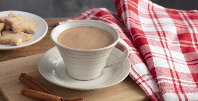 Fotografia em tons de vermelho e marrom de uma bancada, ao centro uma xícara com chocolate quente saindo fumaça e um pires abaixo. Ao lado pedaços de canela, biscoitinhos e uma pano xadrez vermelho e branco.