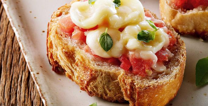 Fotografia em tons de branco e amarelo, com travessa branca e borda dourada com três bruschettas com tomate, mozarela e folhas de manjericão, sobre mesa de madeira rústica.