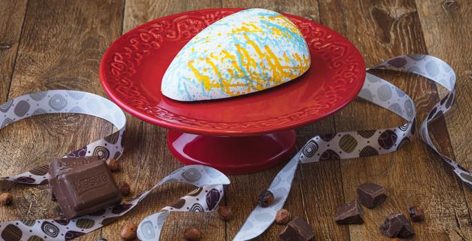 Imagem da receita de uma casca de ovo de páscoa branca, decorada com cores laranja e azul, sobre um prato vermelho, em uma bancada de madeira, decorada com uma fita decorativa, pedaços de chocolate e avelãs