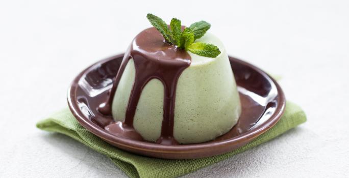 Fotografia de uma mousse de menta com calda de chocolate, com folhas de menta por cima. A mousse está em um prato pequeno na cor marrom, sobre um pano dobrado, na cor verde, que está sobre uma mesa branca.