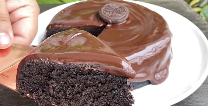 Foto aproximada de um bolo de chocolate com a fatia na frente sobre uma faca. O bolo está decorado com uma calda de chocolate e com um biscoito Negresco no centro.