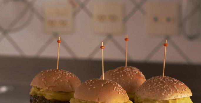 fotografia em tons de cinza e bege de uma bancada marrom vista de frente, contém uma tábua redonda com 4 mini-hambúrgueres com um palito por cima