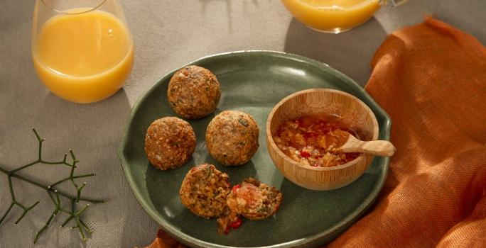 Foto em tons de laranja e verde escuro da receita de bolinho de arroz e aveia servido em 4 porções com uma cumbuca de geleia de pimenta ao lado, tudo sobre um prato de cerâmica verde. Ao lado há um paninho laranja e dois copos de suco de laranja