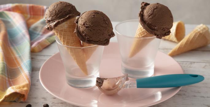 Fotografia em tons de azul e rosa de uma bancada, ao centro um prato rosa com dois copos, dentro deles casquinhas de sorvetes com uma bola do sorvete de café com chocolate. Sobre o prato, um boleador de sorvete com cabo azul.