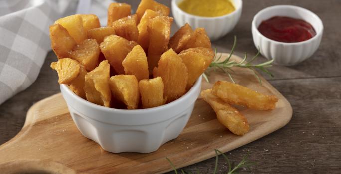 Fotografia em tons de amarelo em uma bancada de madeira escura, uma tábua de madeira, um pote branco fundo com várias mandiocas fritas. Ao fundo, um pano bege xadrez e dois potinhos um com ketchup e um de mostarda.