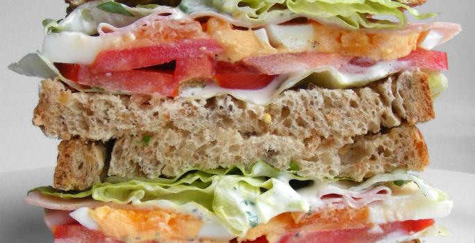 Fotografia de um sanduíche em camadas no pão de forma integral com tomate, repolho, maionese, presuntada e alface. O sanduíche está cortado na metade, sendo uma em cima da outra, que está sobre uma mesa branca.