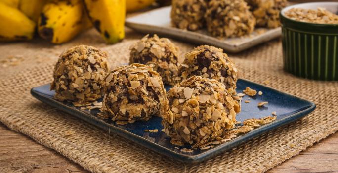 Fotografia em tons de amarelo e azul em uma bancada de madeira, um jogo americano de palha, um recipiente retangular azul com alguns brigadeiros de banana coberto com cereais Neston. Ao fundo, um cacho de banana.