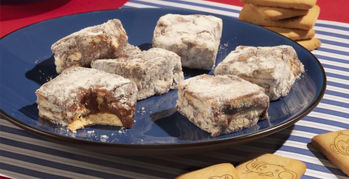 Fotografia em tons de azul e vermelho de uma bancada vermelha com paninho listrado de azul e branco, sobre ele um prato azul escuro com palha italiana de biscoito passatempo. Ao fundo biscoitos passatempo leite espalhados.