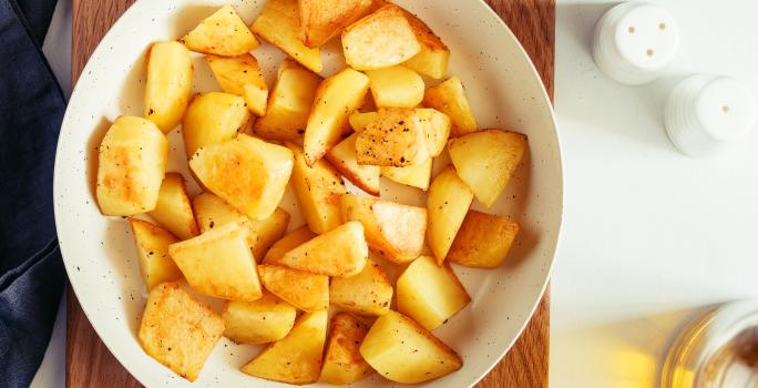 fotografia em tons de amarelo e branco de uma bancada branca vista de cima. Contém uma tábua de madeira marrom, por cima um recipiente redondo branco e dentro batatas picadas.