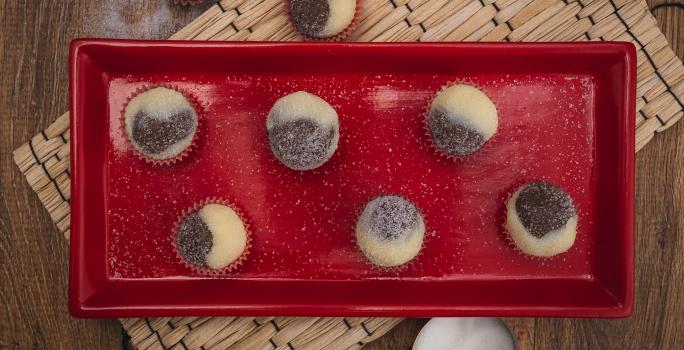 Fotografia em tons de vermelho em uma bancada de madeira escura, um jogo americano de palha, um recipiente retangular vermelho com brigadeiros mesclados de brigadeiro branco e preto em forminhas de vermelho.