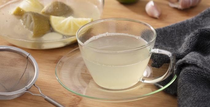 Fotografia em tons de verde em uma bancada de madeira, um pano cinza escuro, um prato de vidro pequeno com uma xícara e o chá de limão com alho dentro dele. Ao fundo, limão e alho espalhado pela mesa.