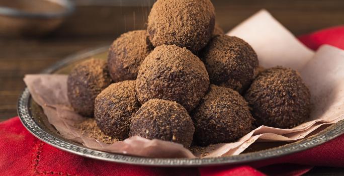 foto em tons de marrom e vermelho de uma bancada marrom vista de frente, contém um pano vermelho, uma bandeja de alumínio com um pano rosa e trufas de chocolate polvilhadas com cacau.