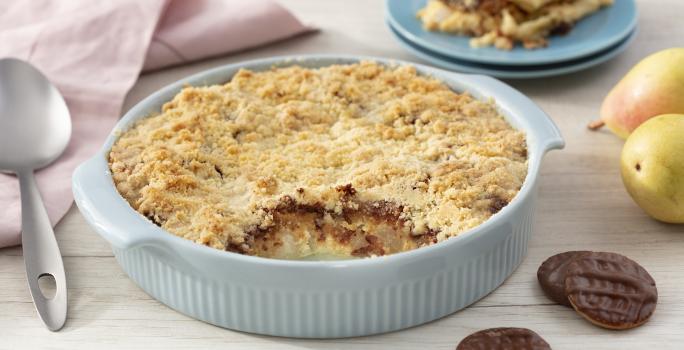 Fotografia em tons de azul em uma bancada de madeira clara, um paninho bege, um recipiente azul claro com o crumble de pera com calipso. Ao fundo, um prato com um pedaço do crumble, peras e calipso espalhados pela mesa.