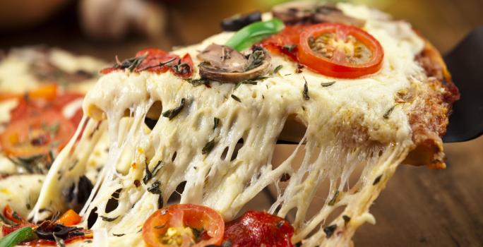 Fotografia de uma pizza com o queijo puxando e bem recheada, como queijo, azeitona, tomates e ricota. A pizza está sobre uma tábua de madeira de toma escuro, e ao fundo, cebola, tomate e champignon.