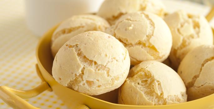 Fotografia em tons de amarelo em uma bancada de madeira esverdeada, um paninho branco com bolinhas amarelas, um recipiente amarelo com vários pães de queijo dentro dele. Ao fundo, uma xícara branca com café.