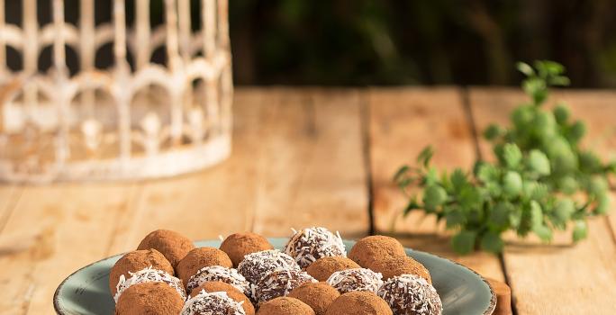 Fotografia em tons de verde em uma bancada de madeira com um prato raso verde ao centro apoiado em uma tábua de madeira e os brigadeiros em cima. Ao lado, brigadeiros espalhados pela bancada.