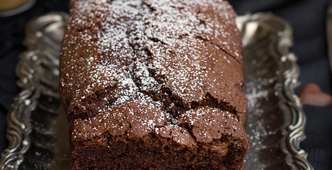 Fotografia em tons de marrom de uma bancada marrom vista de cima, contém uma bandeja de alumínio com um bolo inglês de chocolate com açúcar polvilhado por cima.