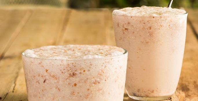 Fotografia em tons de marrom em uma mesa de madeira com dois copos de vidro com o frappé de amêndoas com coco dentro e um canudo de aço inox. Amêndoas espalhadas pela mesa.