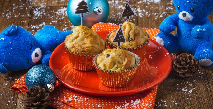 Fotografia em tons de laranja e azul em uma bancada de madeira, um prato laranja com três muffins em cima dele. Ao lado, enfeites e decoração natalina.