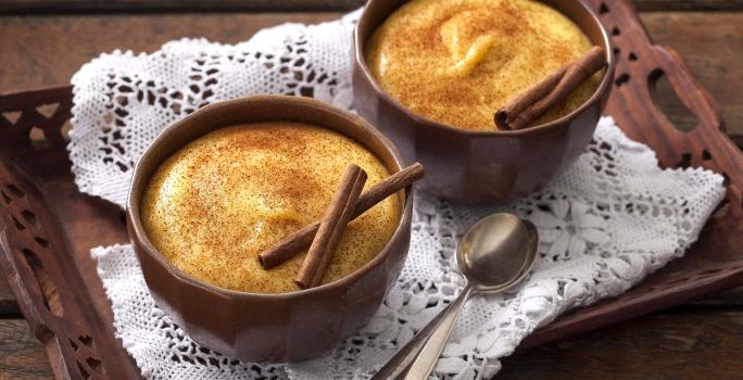 Foto de uma bandeja marrom com uma toalha branca em cima e 2 potes marrons com curau dentro e canela decorando