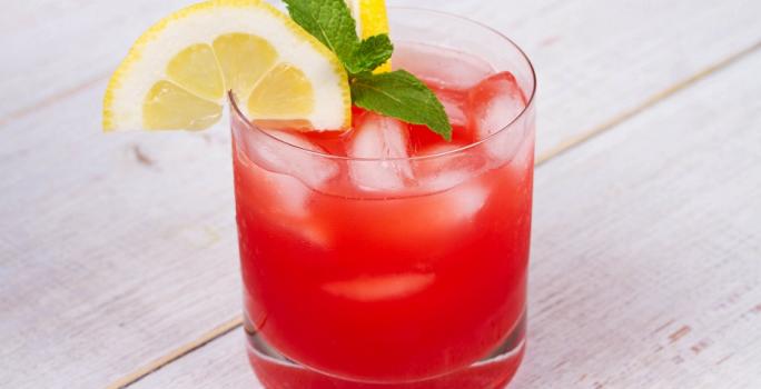 Fotografia em tons de vermelho em uma bancada de madeira de cor branca. Ao centro, um copo contendo a bebida pink lemonade.