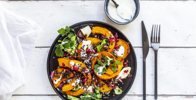 Fotografia vista de cima de uma salada de folhas com molho de laranja, pedaços de abóbora, sementes e um ramo de coentro em um prato raso preto. Ao lado tem talheres e um molho de iogurte em um pote pequeno de vidro azul, com uma colher apoiada.
