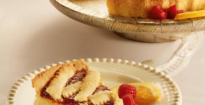 Fotografia em tons de bege em uma mesa de madeira, um guardanapo de pano bege e branco, um prato redondo branco com detalhes bege e uma fatia da crostata com recheio de laranja. Ao fundo, um prato redondo maior com a crostata inteira.