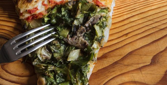 Fotografia em tons de verde em uma bancada de madeira de cor marrom. Ao centro, uma fatia de pizza de escarola com um garfo ao lado.