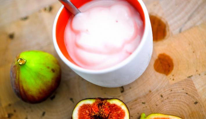 Fotografia de um creme de figo com creme de leite em um recipiente de cerâmica na cor branco e vermelho. Ao redor, dois figos inteiros e mais duas metades do fruto ao lado.