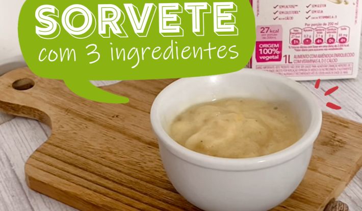 Foto da receita de sorvete de banana servida em um bowl de porcelana branco sobre uma tábua de madeira com amêndoas ao lado direito e bananas em rodelas ao lado esquerdo. Ao fundo, há uma caixa da bebida vegetal de amêndoas nature's heart