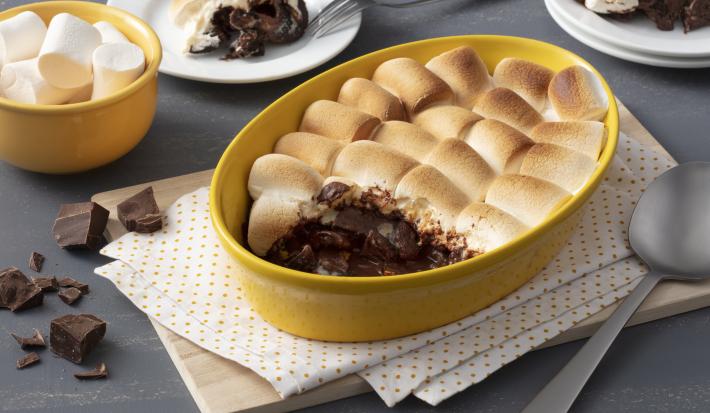 Fotografia em tons de amarelo em uma bancada cinza com um paninho branco de bolinha amarela e um refratário amarelo com o smores de chocolate. Ao fundo, um potinho amarelo com marshmallow e dois pratos com uma fatia do smores em cada um.