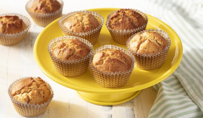 Fotografia em tons de amarelo em uma bancada de madeira branca, um paninho listrado com verde e branco, um prato amarelo alto com vários cupcakes de iogurte com laranja.