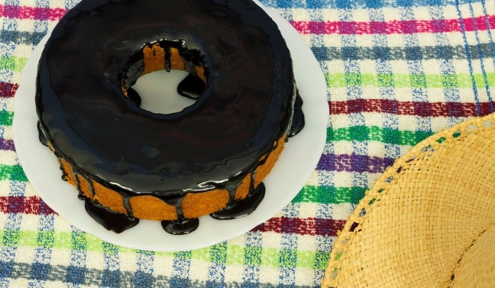 Um bolo de cenoura inteiro com cobertura de chocolate, no formato redondo com furo no meio, está em um prato branco sobre uma toalha colorida xadrez. Um chapéu de palha aparece parcialmente no canto da imagem.