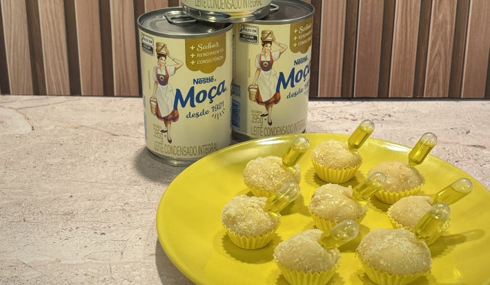 Docinhos cobertos com coco ralado em prato amarelo, com cápsulas decorativas e latas de Leite Moça ao fundo.