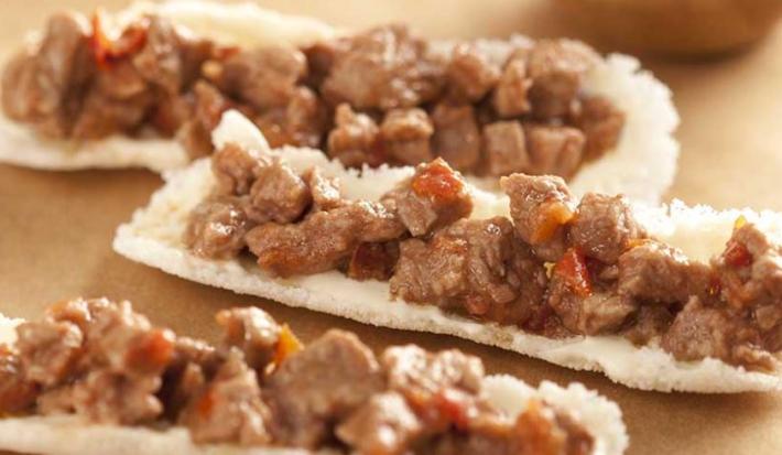 Fotografia em tons de marrom em uma bancada de madeira clara e mini barquinhas feitas com tapioca recheadas com picadinho de carne.