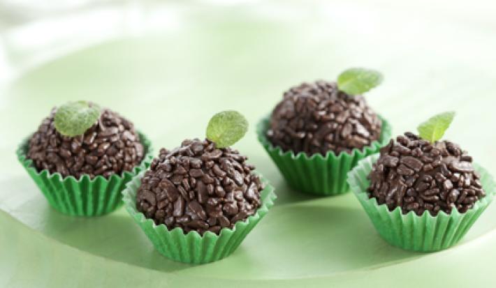 fotografia em tons de verde e marrom, contém 4 brigadeiros com granulados por cima dentro de forminhas verdes e todos tem uma folha de hortelã por cima para decorar