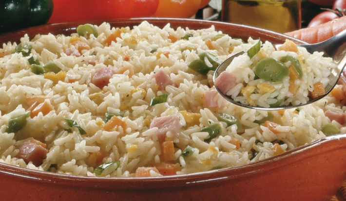 Fotografia em tons de argila e branco, com cumbuca de barro ao centro com arroz e legumes picados sobre guardanapo listrado em tons de argila, ao entorno pimentões, alho, cenoura, tomate-cereja, azeite, pedra vulcânica, tudo sobre bancada de pedra.