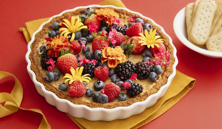 Foto da receita de Torta de Iogurte. Observa-se a torta decorada de frutas vermelhas e flores comestíveis sobre uma superfície vermelha. Ao lado direito, biscoitos inteiros decoram a foto.