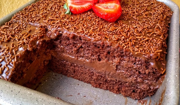 Fotografia em tons claros de uma forma retangular, antiaderente, com o bolo de chocolate, recheado e coberto, finalizado com granulado e pedaços de morango no centro. A foto mostra cerca de dois pedaços do bolo faltando, o que foca na camada do bolo.