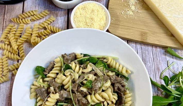 Foto da receita de fusilli com filé mignon e rúcula servida em um prato de porcelana branco sobre uma mesa de madeira. Como decoração, há folhas de rúcula a direita e macarrão a esquerda, além de recipientes com queijo ralado e ingredientes da receita
