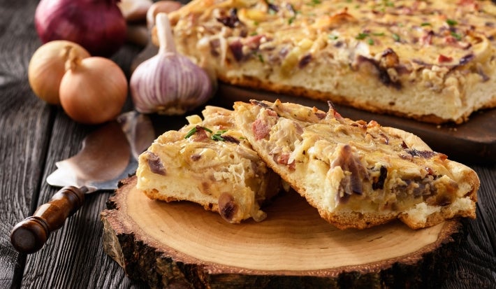 Fotografia de dois pedaços de torta recheada de cebolas sobre um apoio redondo de madeira. Ao fundo tem o restante da torta sobre um apoio de madeira.