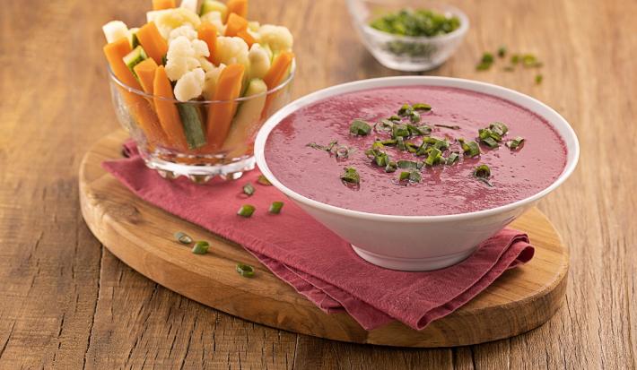 Foto da receita de pasta de beterraba com legumes servida em um bowl de porcelana branca sobre um pano roxo com um recipiente de vidro com legumes variados em fatias ao lado, tudo sobre uma tábua de madeira. Ao fundo, um recipiente de vidro com cebolinha