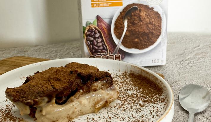 Foto da receita de Bombom de travessa plant-based. Observa-se um pedaço da torta em um prato branco polvilhado de cacau em pó e com com uvas decorando.
