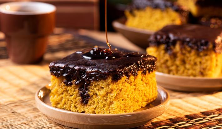 Fotografia em tons de marrom com uma prato pequeno ao centro. Em cima do prato existe uma fatia de um bolo de cenoura de cor alaranjada e com cobertura de chocolate. Ao fundo existe mais fatias desse mesmo bolo.