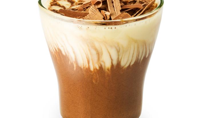 Fotografia em tons de marrom em uma fundo branco, um copo de vidro com a mousse de chocolate e outra mousse de baunilha dentro dele, misturando e formando uma mousse mesclada.