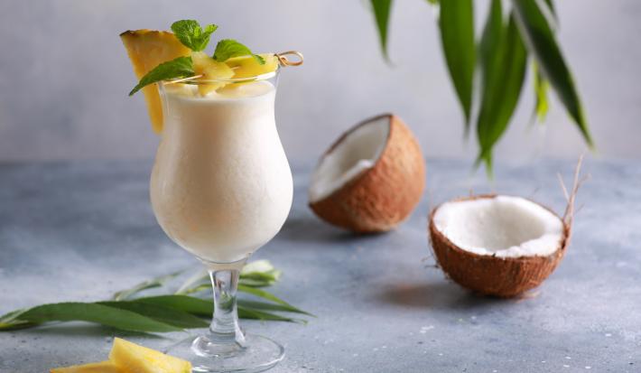 Foto da receita de Piña Colada sem Álcool. Observa-se uma taça grande com a bebida dentro cremosa, uma fatia de abacaxi decorando a borda. Na foto, observa-se um coco seco pela metade.
